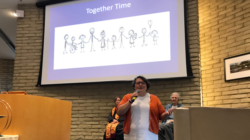 Children's Ministry director speaking in sanctuary; "Together Time" with line drawings of a row of adults and childen is a large projection screen on the wall