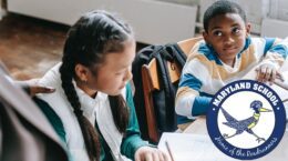 Children sitting at a school table working; one is looking up toward an adult with their hand on the other student's shoulder - overlaid with Maryland School logo