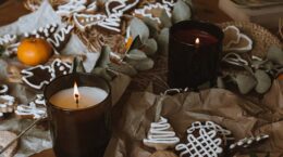 2 lit candles in brown glass holders on table scattered with decorated Christmas cookies