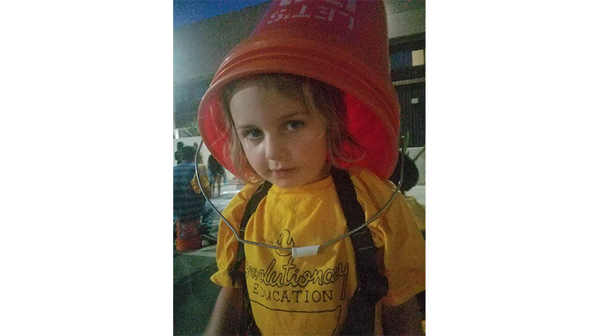 A picture of a kid wearing a yellow t-shirt that says revolutionary education, and has a home depot bucket on her head.