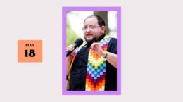 white person with short dark hair and beard, wearing a black shirt and a rainbow quilted stole, holding a microphone