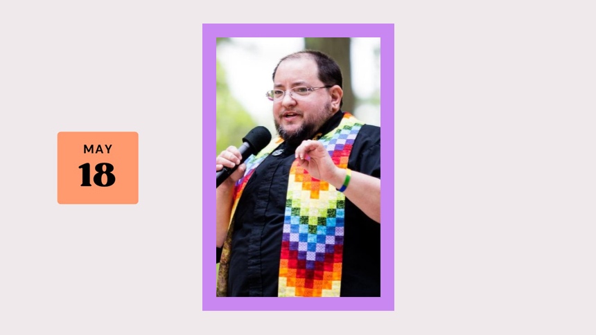 white person with short dark hair and beard, wearing a black shirt and a rainbow quilted stole, holding a microphone